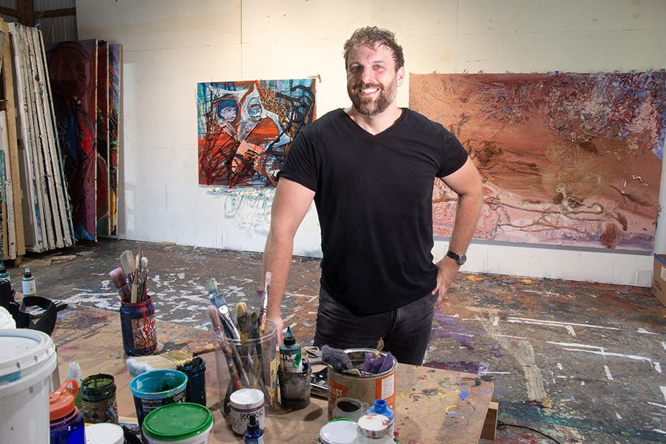 An impossibly handsome and rugged male artist, approximately 40 years old, poses happily in his messy studio with in progress paintings behind him on the wall and a table of paints, brushes, and various tools in front of him.