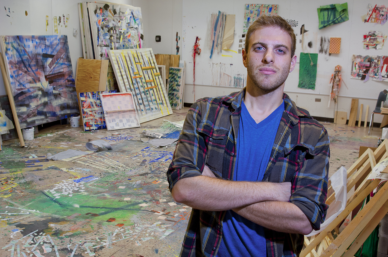 An impossibly handsome baby-faced male artist, approximately 27 years old, poses with attitude in his messy studio with in progress paintings behind him on the wall while at residence at USF.