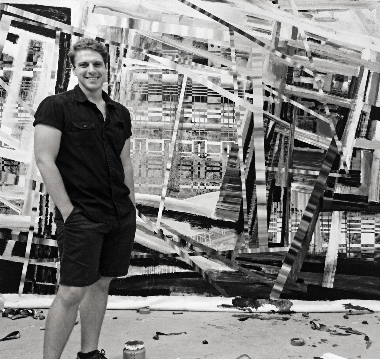 An impossibly handsome male artist, approximately 35 years old, poses happily in his messy studio with in progress paintings behind him on the wall while at residence at VCCA.